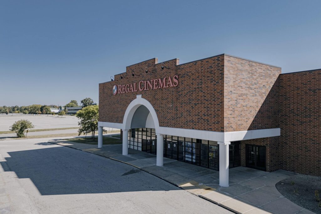 A brick building with the sign 'Regal Cinemas' prominently displayed. The structure features large glass windows and white columns at the entrance, with an expansive empty parking area and greenery visible in the background under a clear blue sky.