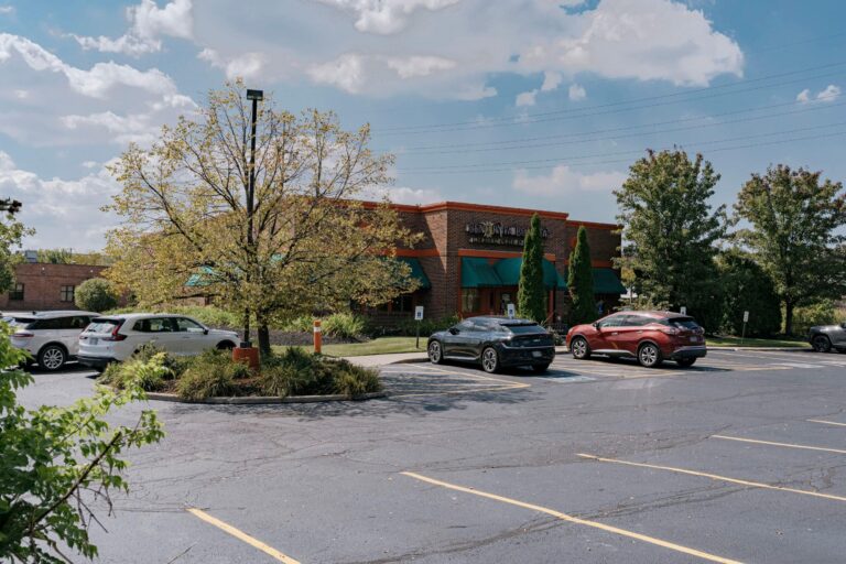 An outdoor view of a restaurant with a brick exterior and green awnings, surrounded by a parking lot with several cars parked. The scene includes a few trees and a cloudy sky in the background, creating a suburban area atmosphere.
