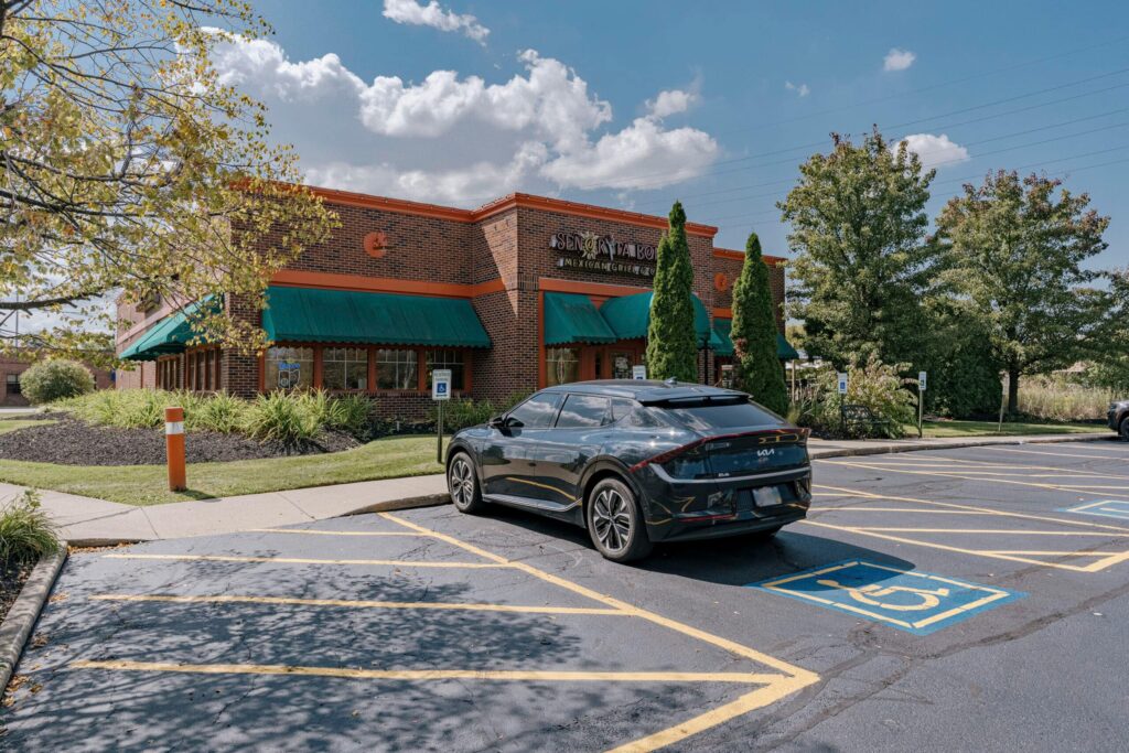 A brick building with green awnings identified as a Mexican grill and cantina. The foreground shows a parking lot with a black car parked in a handicapped parking spot, surrounded by plants and trees under a partially cloudy sky.