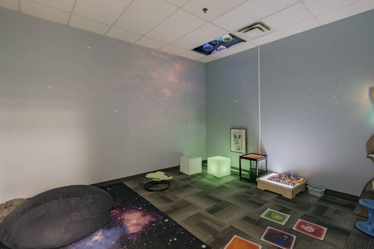 A sensory room featuring a light gray wall with projected stars and lights, colorful ceiling lamps, a galaxy-themed carpet with a bean bag, cube seating with soft lighting, a table with colorful buttons, and various sensory mats on the carpeted floor.