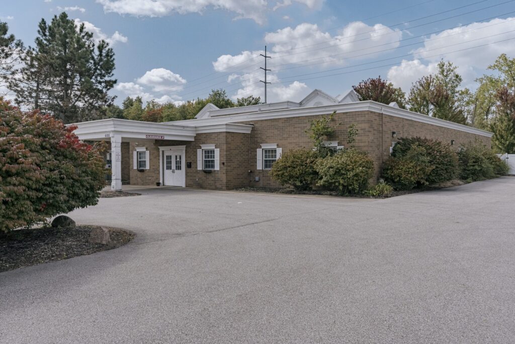 A single-story brick building with white trim and double doors, surrounded by bushes and trees. The sky is partly cloudy, and the building is adjacent to a paved driveway with a clearance sign visible.