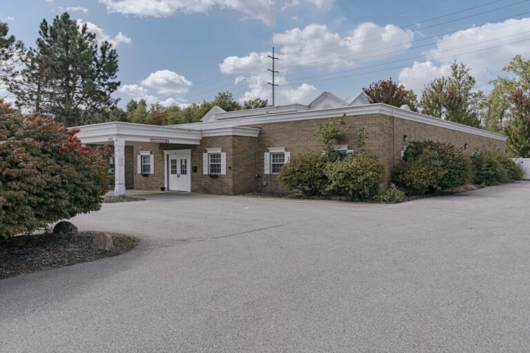 A single-story brick building with white trim and double doors, surrounded by bushes and trees. The sky is partly cloudy, and the building is adjacent to a paved driveway with a clearance sign visible.