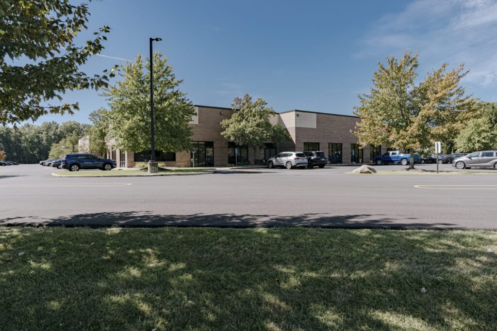 A suburban one-story brick office building with large windows, surrounded by a parking lot filled with several cars. The scene includes green grass in the foreground and trees lining the perimeter, under a clear blue sky.