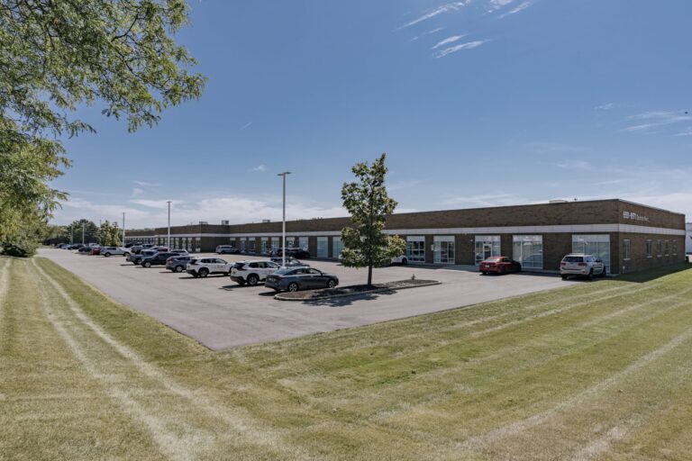 A single-story brick office building with large glass windows, situated under a clear blue sky. The building is surrounded by a neatly maintained parking lot with cars parked in rows. In the foreground, there is a grassy area with trees, giving the scene a suburban feel.