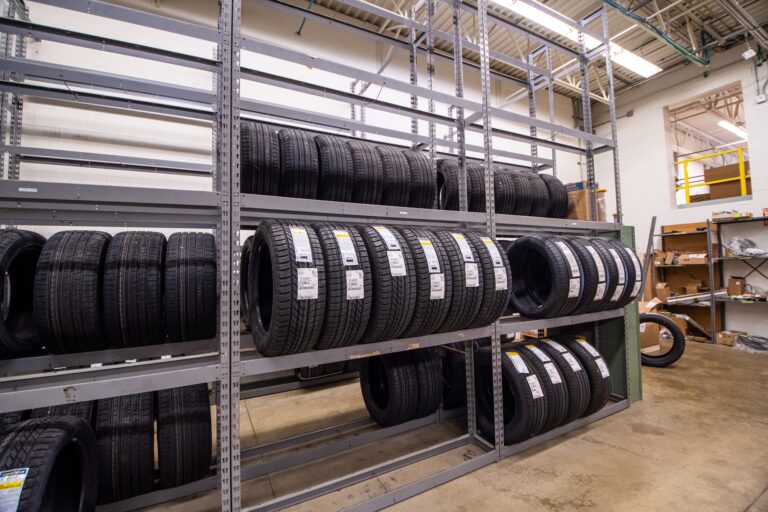 A warehouse interior filled with black tires arranged on metal shelving units. The floor is concrete and the space has high ceilings with metal beams. Some tires have tags with information labels attached, and there are other storage racks with various items in the background.