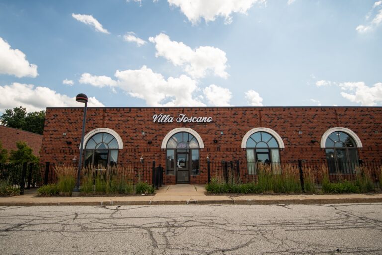 A brick building with arched windows and a sign reading 'Villa Toscano'. The entrance features a glass door with decorative plants on either side, and a clear blue sky overhead.