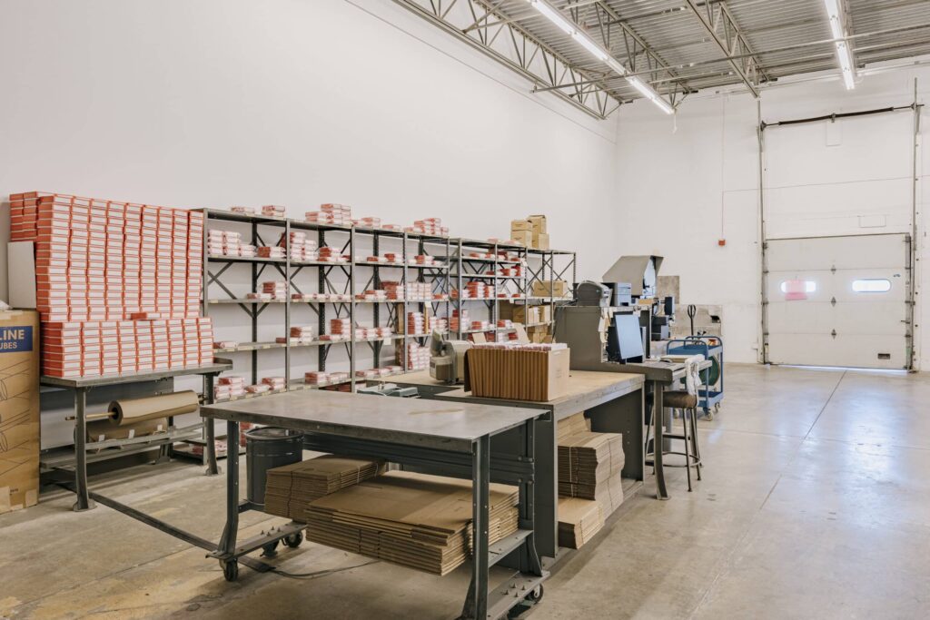 Industrial warehouse space with shelves filled with red and white boxes, large tables stacked with cardboard packages, and rolls of paper. The room has industrial lighting, a concrete floor, and a large roll-up door.
