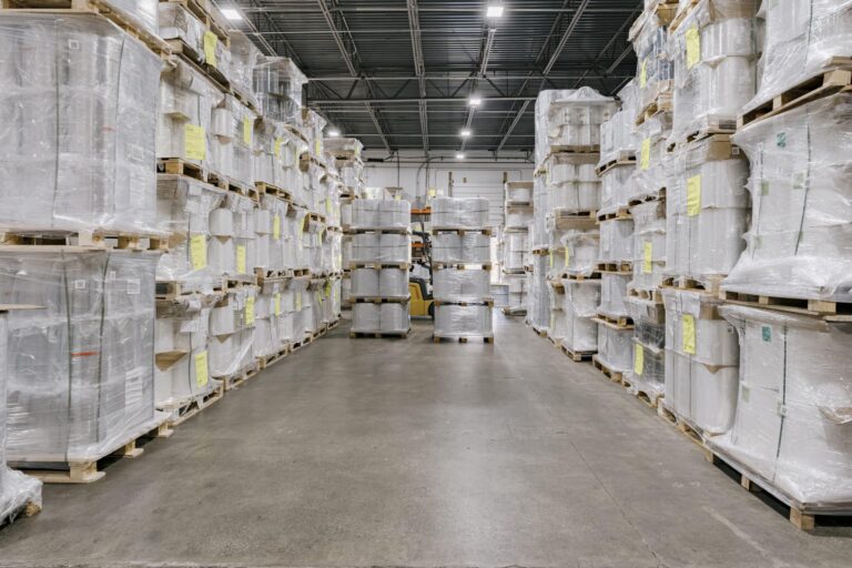 A spacious warehouse interior with stacks of pallets wrapped in clear plastic, organized in rows on both sides of a wide aisle. The pallets are filled with boxed goods, and there is a forklift visible in the distance, indicating ongoing operations.