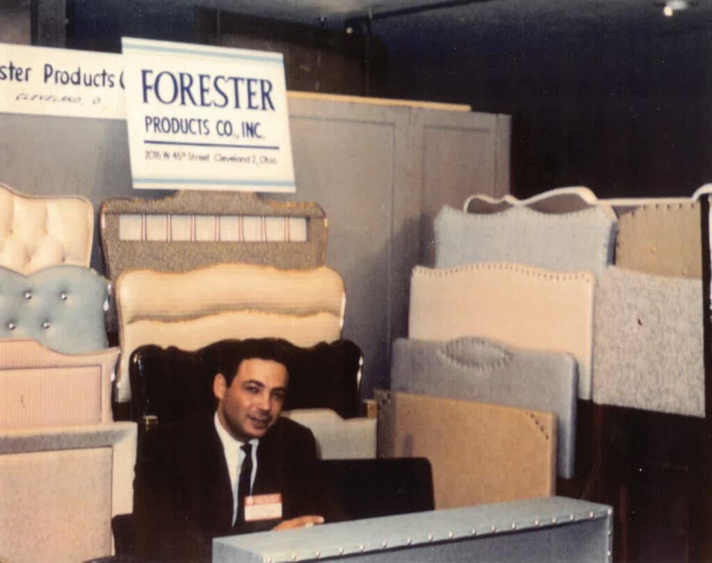 A man in a suit is sitting at a booth for Forester Products Co., Inc. The booth displays various upholstered headboards in different designs and colors. A sign above the booth shows the company's name and address in Cleveland, Ohio.
